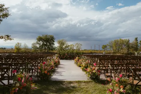 An outdoor wedding ceremony setup featuring chairs arranged along a floral-lined aisle under a cloudy sky.