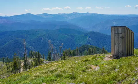 A wooden outhouse sits on a grassy mountain ridge overlooking vast distant ranges.