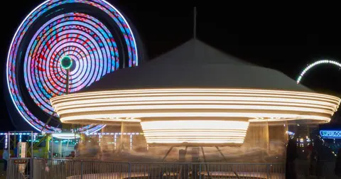 A brightly lit carousel spinning at night with a large Ferris wheel in the background.