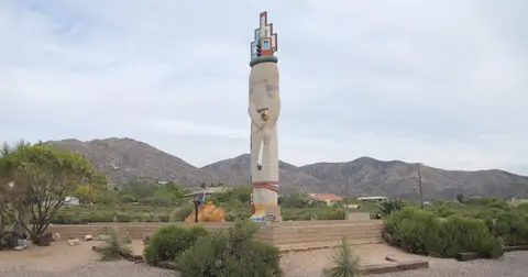 A tall statue stands outdoors in a desert landscape with mountains in the background.