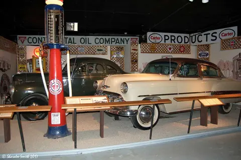 A vintage gas pump and two classic cars are displayed inside a museum.