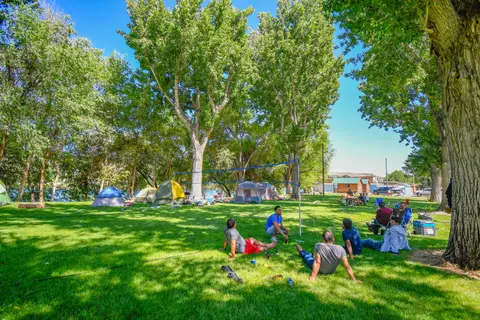 A group of people sitting on the grass near a volleyball net at a campground with tents and trees.