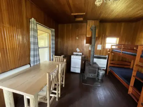An interior view of a rustic wooden cabin featuring a dining table, a wood-burning stove, and bunk beds.