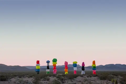 A row of brightly painted stacked boulders stands in a desert landscape against a clear sky.
