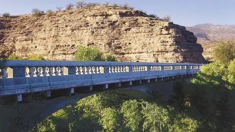 A concrete bridge with a balustrade crosses a landscape with lush trees and a rocky hill in the background.