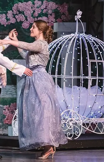 A woman in a blue gown dances near a decorated carriage stage prop.