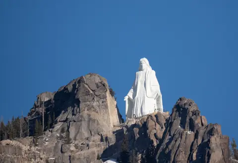 A large white statue of a figure stands on a rocky mountain ridge under a clear blue sky.