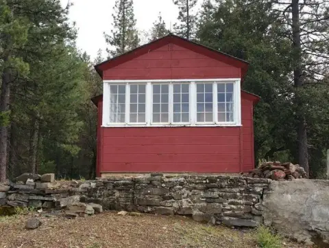 A red wooden building with white-trimmed windows stands behind a stone wall in a forest.