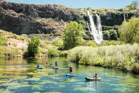 People enjoy paddleboarding and kayaking in a clear river with a waterfall and cliffs in the background.