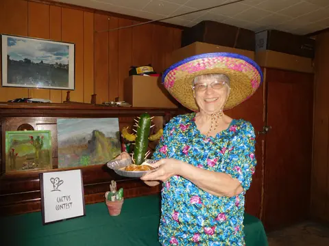 A smiling woman wearing a sombrero holds a pickle decorated like a cactus.