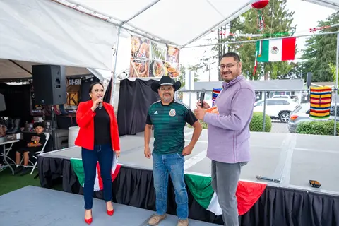 Three people stand on a stage under a tent at an outdoor event with Mexican flags.