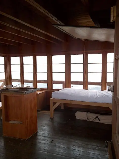 The interior of a wooden cabin or fire lookout tower featuring a bed and a view through multiple windows.