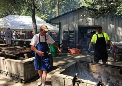 Two men cooking meat on large outdoor grills at an event.