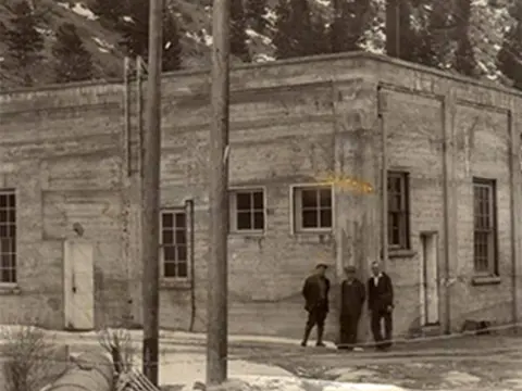 Historic photo showing the exterior of a concrete building with three people standing near the corner.