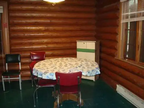 An interior shot of a room with log walls, a table with chairs, and a small cabinet.