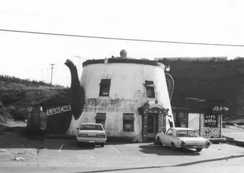 Black and white exterior photo of a teapot-shaped building with parked cars in front.