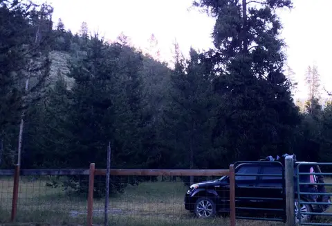 A car parked by a fence with dense trees and a hill in the background.