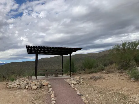 A shaded picnic table sits along a desert path under a cloudy sky.