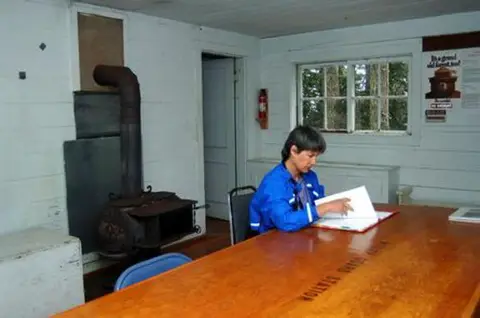 A person sits at a wooden table inside a rustic room with a wood-burning stove.