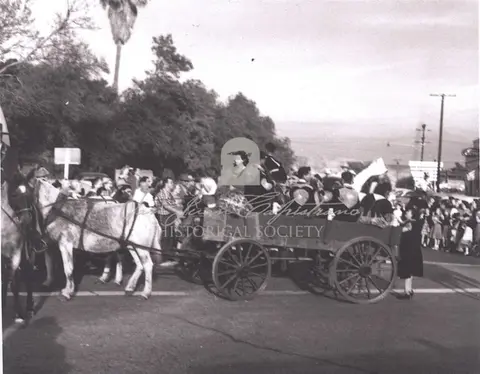 Black and white historic photo of a horse-drawn wagon in a parade with onlookers.