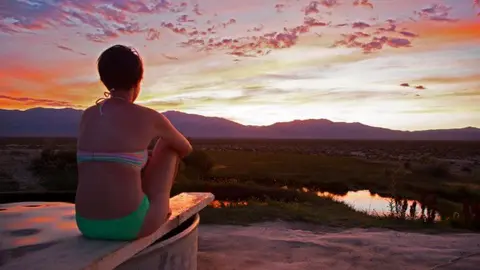 A person sits on the edge of a tub overlooking a desert landscape and mountains at sunset.