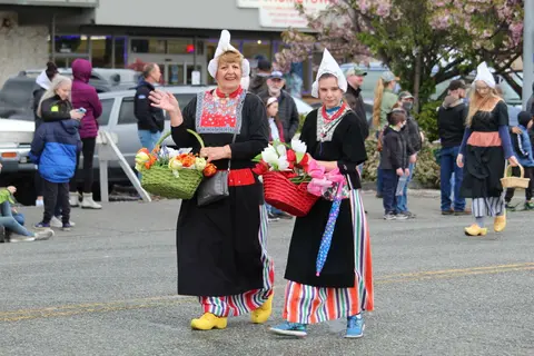 Two women in traditional Dutch attire walking in a parade holding baskets of flowers.