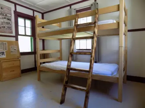 A wooden bunk bed stands inside an interior room with a window and a bulletin board.