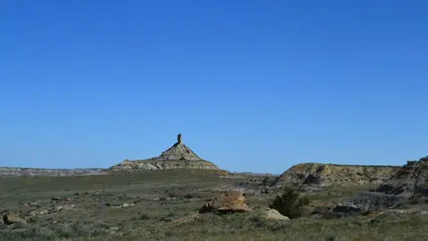 A tall rock formation rises above a dry landscape under a clear blue sky.