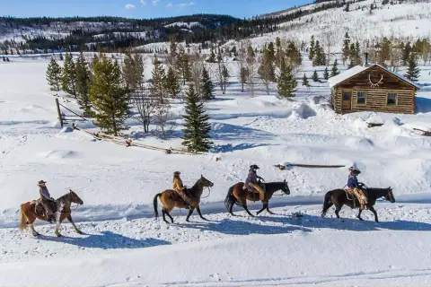 Four people ride horses on a snowy landscape past a log cabin.