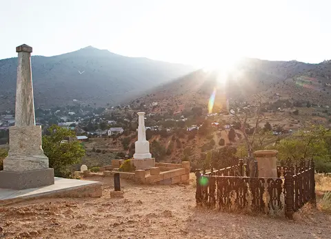 Monuments and grave markers stand on a hillside overlooking a town with mountains in the background.