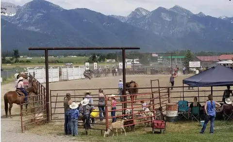 People and horses gather at a rodeo arena with mountains in the background.