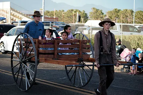 People in period costume pull and ride in a wooden wagon.