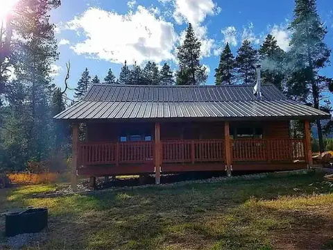 A wooden cabin with a metal roof sits in a clearing surrounded by trees.