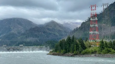 A large dam and transmission towers sit near a river beneath cloudy mountains.
