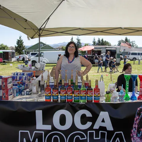A woman stands behind a refreshment stand outdoors.