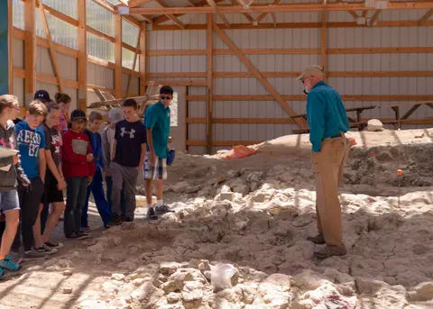 A guide speaks to a group of young people inside a large wooden pavilion over an archaeological dig site.