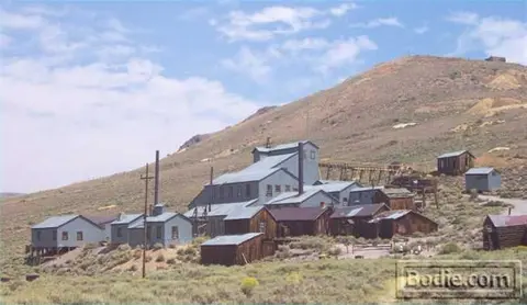 Old wooden buildings and industrial structures sit on a barren hillside beneath a cloudy sky.