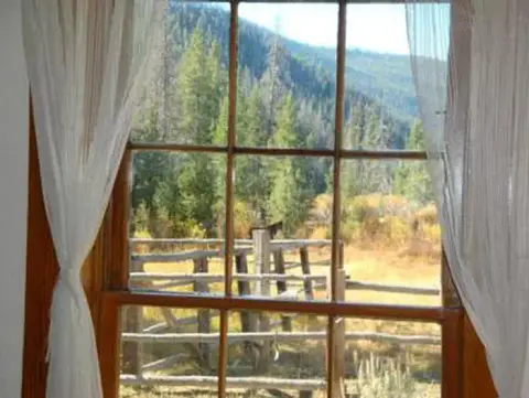 A mountain landscape and wooden fence viewed through a window with white curtains.