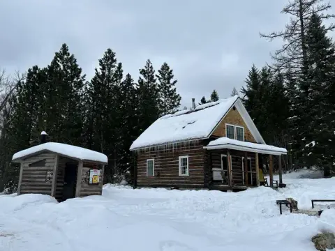A snow-covered log cabin and outhouse surrounded by pine trees in winter.