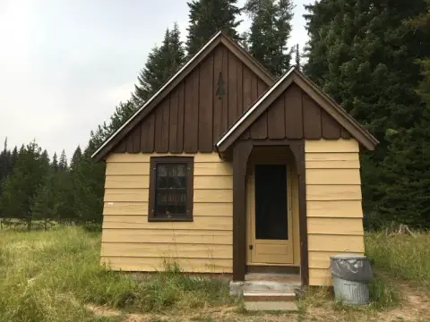 A small cabin stands surrounded by grassy fields and tall trees.