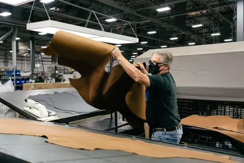 A worker handles a large sheet of brown leather in an industrial workshop.