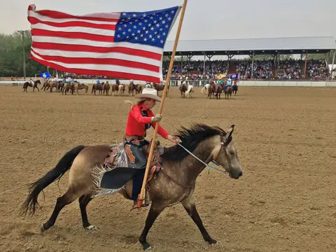 A cowgirl rides a horse while carrying an American flag in an arena.