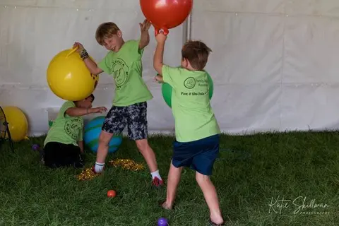 Three children play with large colorful bouncy balls in a grassy area inside a tent.