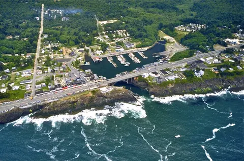 An aerial view shows a coastal town with a marina, bridge, and crashing waves.