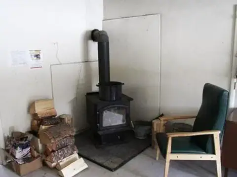 A wood-burning stove sits in the corner of a room near a stack of firewood and an armchair.