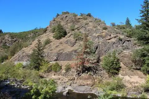 A rocky hill covered in sparse vegetation with a stream at its base under a blue sky.