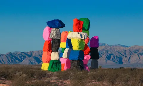 A large sculpture made of stacked, brightly painted boulders stands in a desert landscape.
