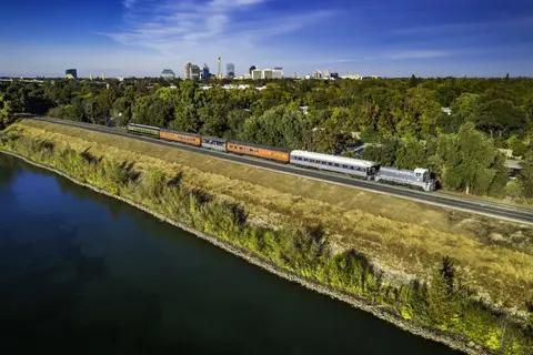 A train travels along a riverbank with a city skyline in the background.