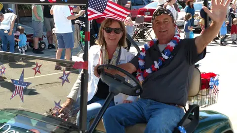 A man and woman wearing patriotic accessories sit in a golf cart decorated for a parade.