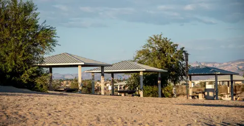 A row of covered picnic shelters stands outdoors near trees and a sandy area.